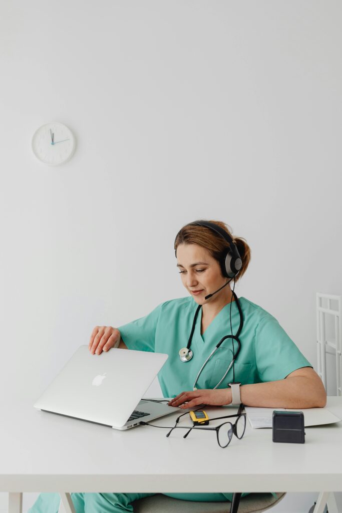 A female doctor in scrubs conducts an online consultation using a laptop.